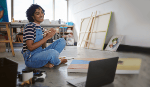 Woman sitting in front of painitng canvases in blue overalls 